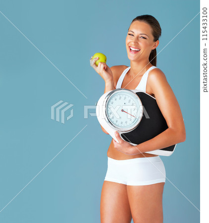 Woman, scale and apple for weight loss, portrait and nutrition on blue studio background. Healthy, wellness and excited for diet, mockup space and winner with victory, organic food or body positivity 115433100