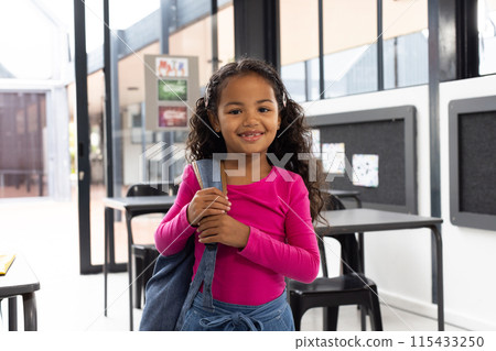 Biracial girl with curly hair smiles, holding a backpack in a school classroom Biracial girl with curly hair smiles, holding a backpack in a school classroom 115433250