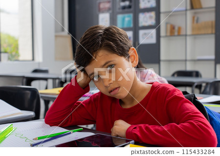 Biracial boy with brown hair looks sad in a school classroom setting 115433284