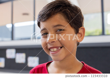 Biracial boy with a bright smile, wearing a red shirt in a school classroom 115433310