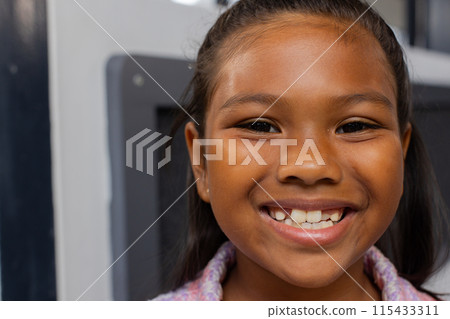 Biracial girl with a bright smile, wearing a colorful top in a school classroom 115433311
