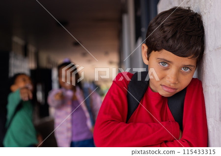 Biracial boy leans against a white wall, looking pensive, a victim of bullying in school 115433315