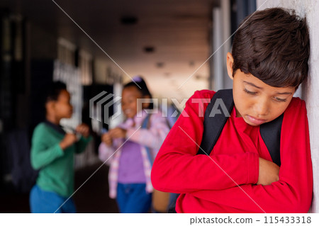 Biracial boy in a red shirt looks upset, leaning against a wall in school, experiencing bullying 115433318