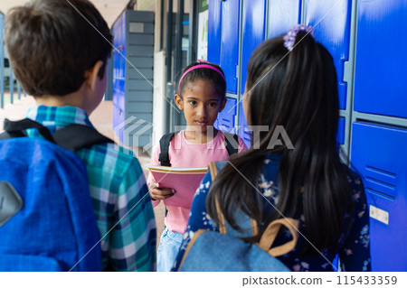 Biracial girl with pink headband talks to her peers by blue school lockers, holding a notebook 115433359