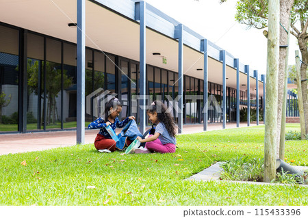 Two biracial girls are sitting on the grass in school, eating lunch with copy space 115433396