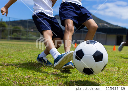 Children in sports attire are playing soccer on a grassy field during the day in school 115433489