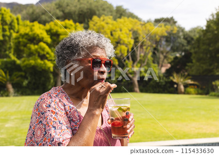 Senior biracial woman enjoys a drink outdoors, wearing red sunglasses and a patterned blouse 115433630