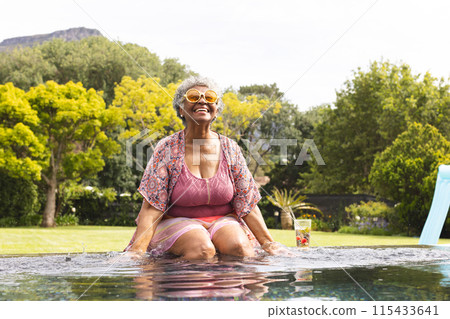 Senior biracial woman enjoys a sunny day in the pool, wearing a floral top and sunglasses Senior biracial woman enjoys a sunny day in the pool, wearing a floral top and sunglasses 115433641