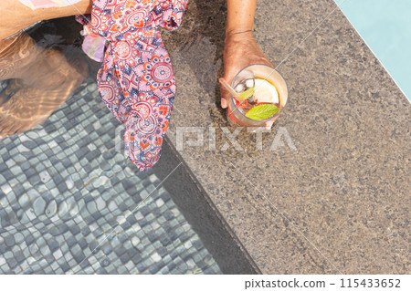 Senior biracial woman enjoys a poolside retreat with copy space, her hand holding a refreshing drink 115433652