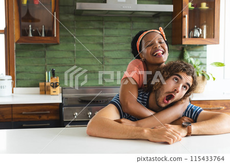 Diverse couple young African American woman embraces a young Caucasian man from behind in a kitchen Diverse couple young African American woman embraces a young Caucasian man from behind in a kitchen 115433764