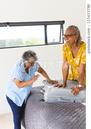 Senior African American woman and senior biracial woman share a joyful moment over a suitcase with c 115433796