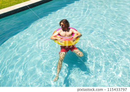 Young Caucasian man floats in a pool with a colorful ring, wearing sunglasses and red shorts Young Caucasian man floats in a pool with a colorful ring, wearing sunglasses and red shorts 115433797