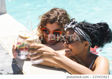 Diverse couple enjoying drinks by the pool, the woman's hair adorned with a headband Diverse couple enjoying drinks by the pool, the woman's hair adorned with a headband 115433821