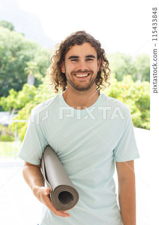 Young Caucasian man with curly hair holds a yoga mat, smiling outdoors 115433848
