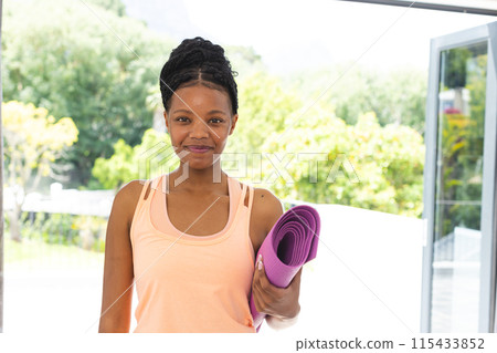 Young African American woman holding a purple yoga mat, wearing a light orange tank top 115433852
