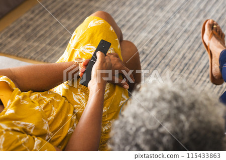 Senior African American woman in a yellow dress holds a phone, seated indoors at home Senior African American woman in a yellow dress holds a phone, seated indoors at home 115433863