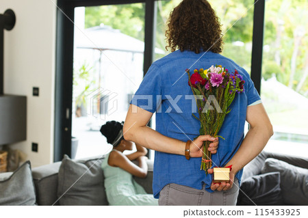 Diverse couple: Young Caucasian man surprises African American woman with flowers and a gift box 115433925