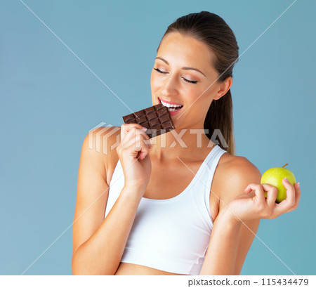 Apple, woman and chocolate choice in studio for health, food and diet plan in isolated blue background. Smile, green fruit and face of young vegetarian person for detox, vegan or decision 115434479