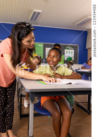 Asian teacher assists a smiling African American girl with schoolwork in a school classroom 115434488