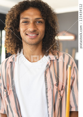 Young biracial man with curly hair smiles warmly, holding a pool cue 115434512