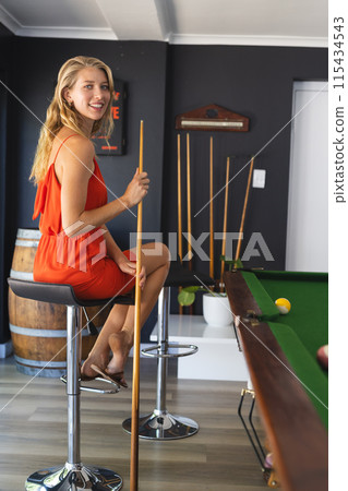 Young Caucasian woman in an orange dress enjoys a game of pool, holding a cue stick with copy space Young Caucasian woman in an orange dress enjoys a game of pool, holding a cue stick with copy space 115434543