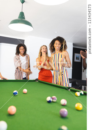 Young biracial man and two young Caucasian women enjoy a game of pool 115434573