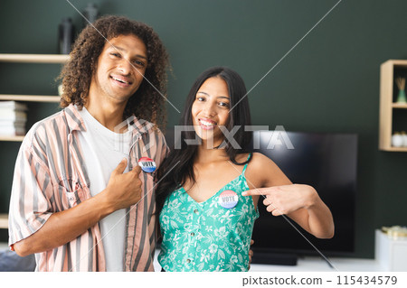Biracial couple proudly displaying "I Voted" stickers, standing in a home setting 115434579