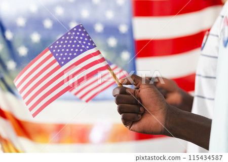 African American man's hand holding a small American flag, with a larger flag in the background African American man's hand holding a small American flag, with a larger flag in the background 115434587