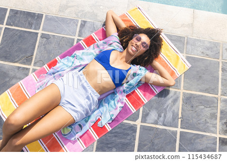 Biracial woman enjoys the sun by a pool, wearing a blue bikini top and sunglasses 115434687