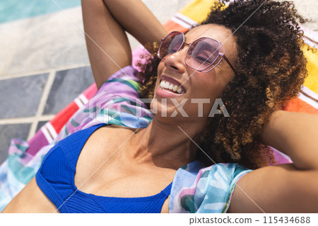 Biracial woman enjoys the sun by the pool, wearing pink sunglasses and a blue swimsuit Biracial woman enjoys the sun by the pool, wearing pink sunglasses and a blue swimsuit 115434688