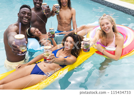 Diverse group of friends enjoying a pool party, toasting drinks 115434733