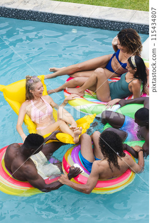 Diverse group of friends enjoy a pool party, lounging on colorful floaties 115434807