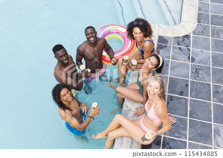 Diverse group of friends enjoy a pool party, holding tropical drinks 115434885