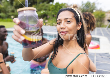 Young biracial woman in a green swimsuit holds out a fruit-infused drink, smiling at a pool party 115434888
