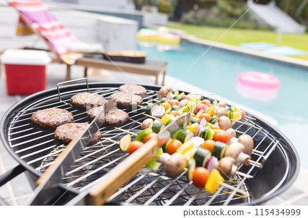 Burgers and vegetable skewers are grilling by a poolside 115434999