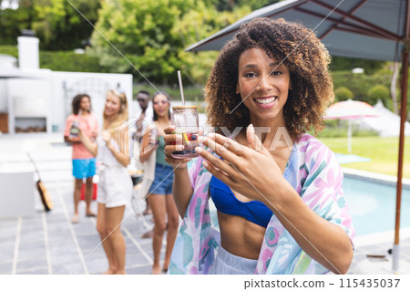 Young biracial woman smiling, holding a drink at a pool party 115435037