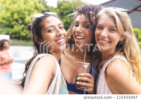 Three smiling diverse female friends take a selfie, one holding a drink 115435047