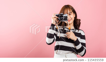 Attractive energetic happy Asian portrait beautiful young woman smiling photographer taking a picture and looking viewfinder on retro vintage photo camera ready to shoot isolated on pink background 115435559