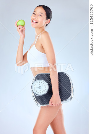 Portrait, scale and happy woman with apple in studio for health, diet and weight loss detox on grey background. Nutrition, wellness and face of girl model with fruit or organic, vegan or gut health 115435769