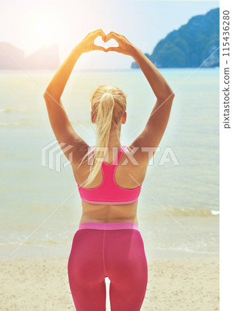 Fitness, yoga and heart hands with woman on beach for wellness, mental health or exercise in nature. Environment, spiritual peace and athlete back doing warm up, zen wellbeing and ocean workout 115436280