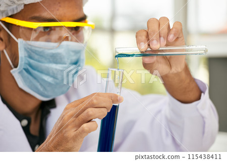 Close-up of scientist wearing safety goggles and mask, carefully pouring blue liquid from test tube into graduated cylinder in laboratory setting 115438411