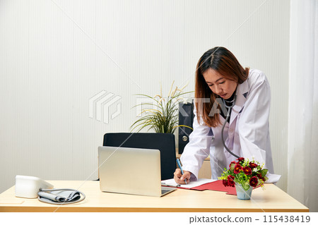 Asian female doctor in white coat writing patient notes on clipboard while standing at desk with laptop and medical equipment, emphasizing professionalism and thoroughness 115438419
