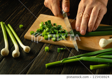 The cook cuts green young garlic on a wooden cutting board with a knife for preparing vegetarian food. Peasant dish The cook cuts green young garlic on a wooden cutting board with a knife for preparing vegetarian food. Peasant dish 115438924