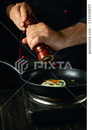 The chef uses a hand mill to add pepper to a hot frying pan with eggs. Low key concept of preparing breakfast with egg in restaurant kitchen 115438925