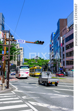 Kumamoto Castle stands out against the blue sky, and trams and road traffic run through the downtown area (Torimachi-suji, Kumamoto City) 115439087