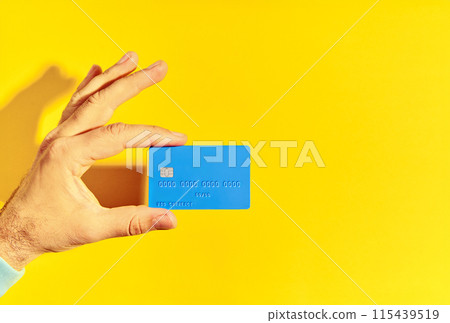 Male hand holds blue plastic bank card over yellow background with copy space Male hand holds blue plastic bank card over yellow background with copy space 115439519