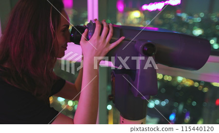Woman looking in telescope at night city. Tourist enjoys Bangkok Baiyoke Tower Sky viewpoint. Colorful cityscape center downtown, night lights view from top floor. Travel, tourism 115440120