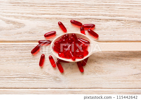 Vitamin capsules in a spoon on a colored background. Pills served as a healthy meal. Red soft gel vitamin supplement capsules on spoon 115442642