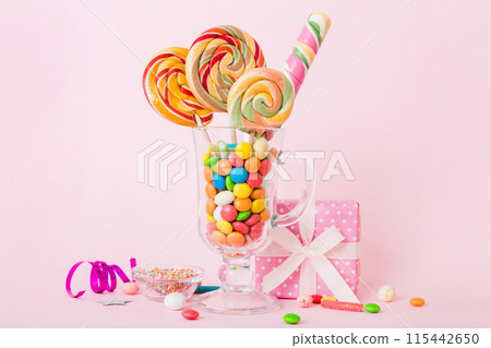 Colorful candies in cup on table on light background background. Large swirled lollipops. Creative concept of a jar full of delicious sweets from the candy store Colorful candies in cup on table on light background background. Large swirled lollipops. Creative concept of a jar full of delicious sweets from the candy store 115442650