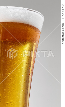 Close-up of pint glass filled with golden, craft beer topped with frothy foam against grey studio background. Close-up of pint glass filled with golden, craft beer topped with frothy foam against grey studio background. 115443735